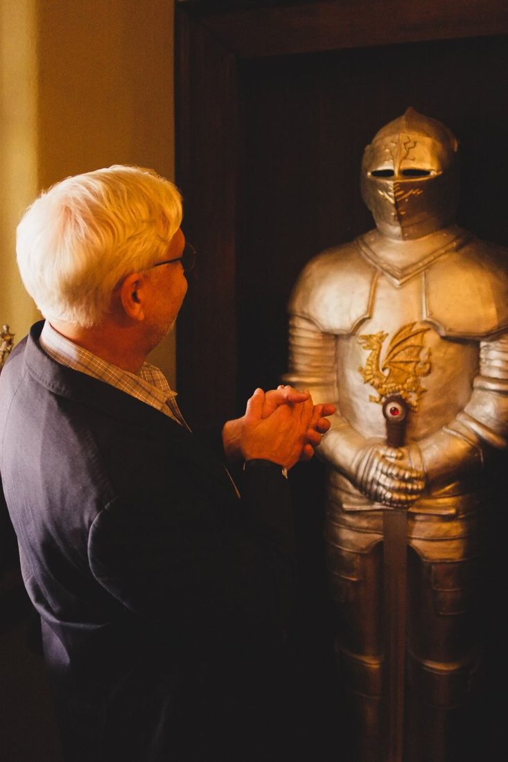 Gabriel Ramsey in a suit, standing beside a suit of armor.